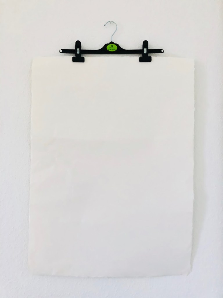 Eine Fotografie der Installation Spaceholder. Zu sehen ist ein Aquarellpapier, das an einem Bügel befestigt an der Wand hängt.